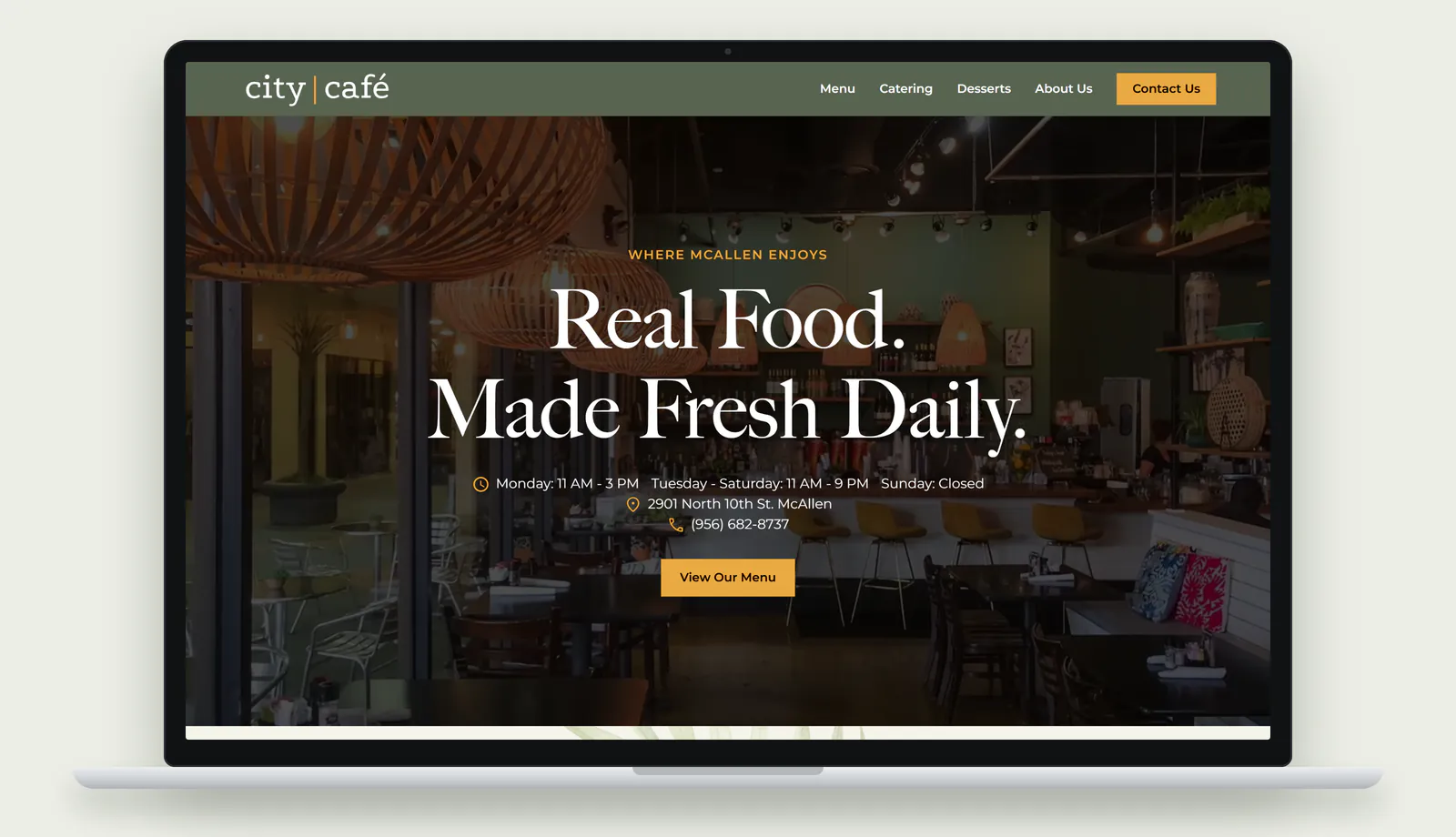 The new City Café & Catering homepage on a laptop: a sage green navigation bar with the city café logo, links to Menu, Catering, Desserts and About Us, and a gold Contact Us button, over a darkened photo of the dining room with woven pendant lights. A gold line reading Where McAllen Enjoys sits above the headline Real Food, Made Fresh Daily, followed by the hours, the North 10th Street address, the phone number and a gold View Our Menu button.